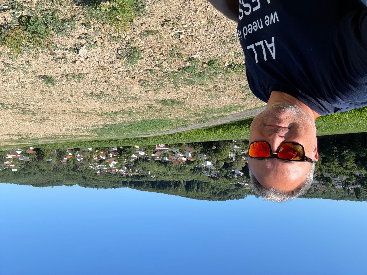 A man wearing a black shirt with white text and sunglasses is looking down at a valley with houses and trees.