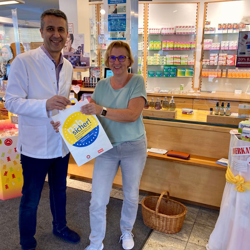 Ein Mann und eine Frau stehen in einer Apotheke, die Frau hält einen Beutel mit der Aufschrift 'sicher!'. Der Mann lächelt und trägt einen weißen Laborkittel. Hinter ihnen ist eine Theke mit verschiedenen Produkten zu sehen.