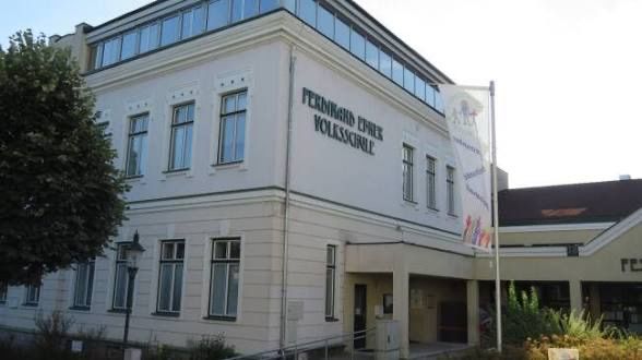 Ein weißes Gebäude mit mehreren Fenstern und einem Fahnenmast vorn. Das Gebäude hat ein Namensschild, das 'Ferdinand-Ehrler-Volksschule' lautet.