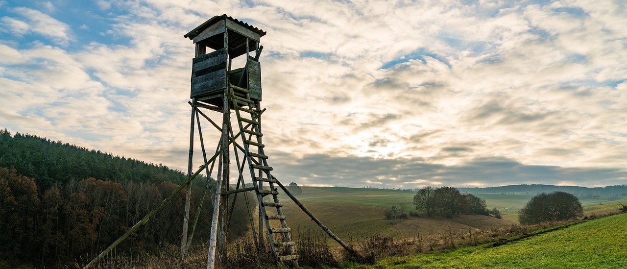 Ein alter Holzaussichtsturm steht auf einem grasigen Hügel mit Blick auf die umliegende Landschaft.