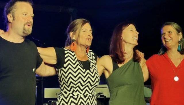 Vier Menschen stehen gemeinsam auf einer Bühne, lächeln und scheinen für ein Foto zu posieren. Der Mann links hat einen Bart und die Frau in der Mitte rote Ohrringe. Hinter ihnen stehen ein Klavier und andere Musikinstrumente.