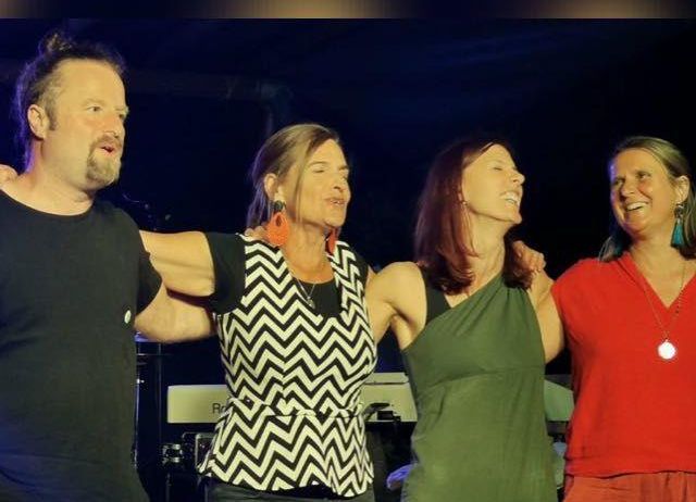 Vier Menschen stehen gemeinsam auf einer Bühne, lächeln und scheinen für ein Foto zu posieren. Der Mann links hat einen Bart und die Frau in der Mitte rote Ohrringe. Hinter ihnen stehen ein Klavier und andere Musikinstrumente.