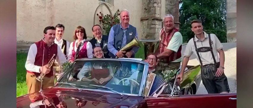 Eine Gruppe Musiker in traditioneller Kleidung posiert mit einem Oldtimer vor einem historischen Gebäude.