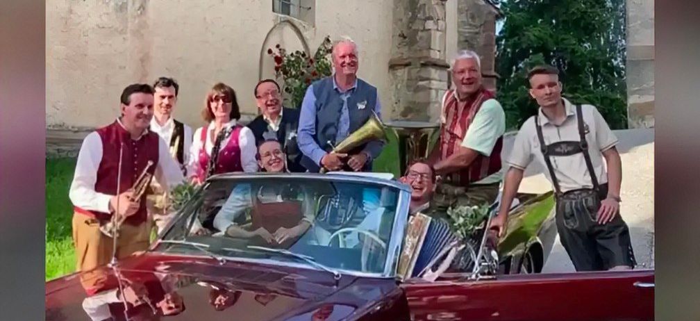 Eine Gruppe Musiker in traditioneller Kleidung posiert mit einem Oldtimer vor einem historischen Gebäude.