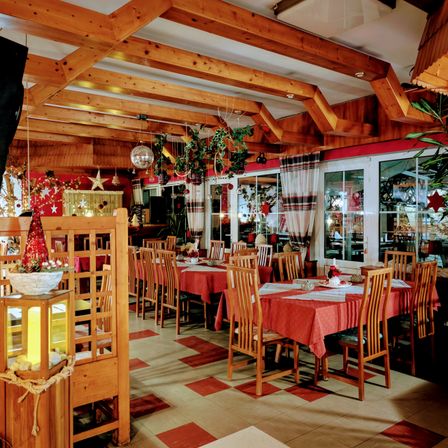 Ein Restaurant mit Holzbalken, einem karierten Boden und Weihnachtsdekorationen. Tische sind mit roten Tischdecken und Blumengestecken gedeckt. Fenster und Türen lassen natürliches Licht herein.