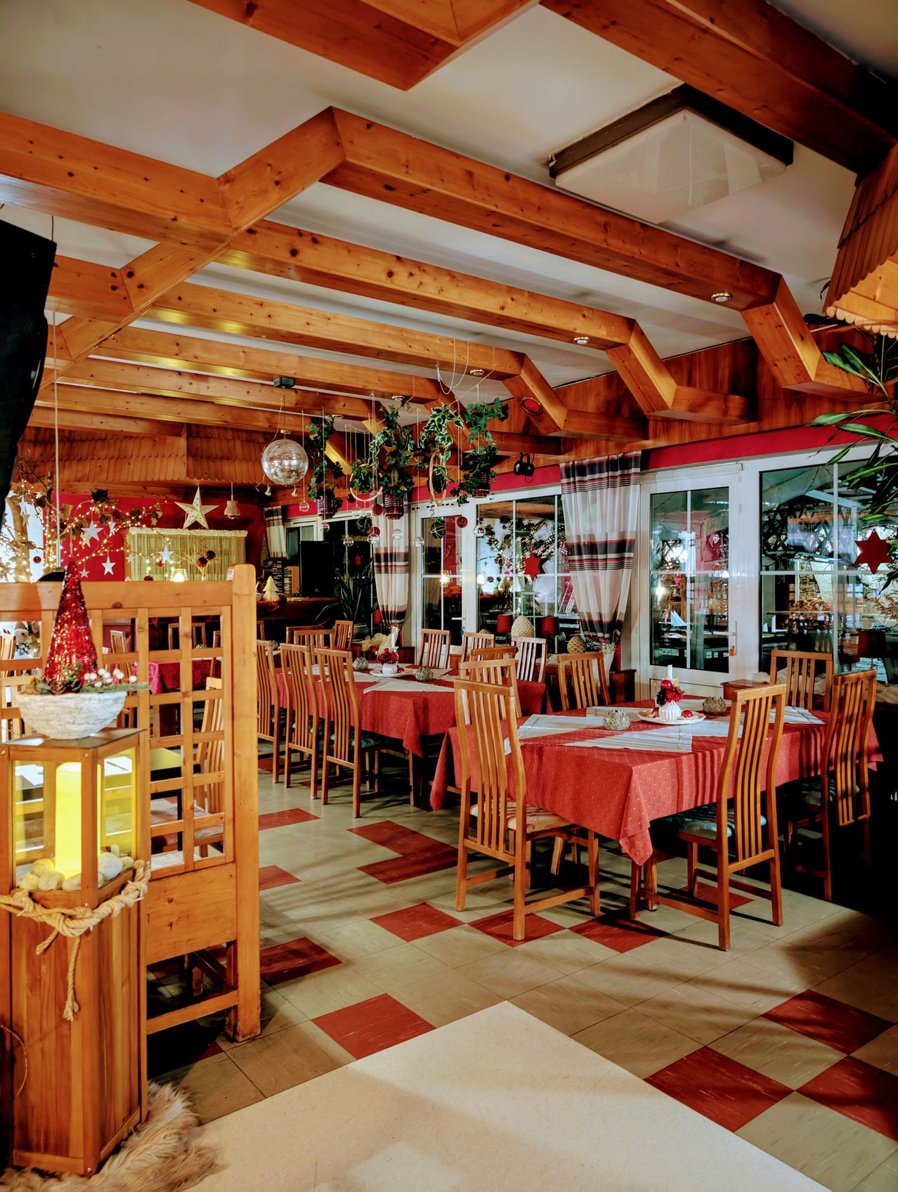 Ein Restaurant mit Holzbalken, einem karierten Boden und Weihnachtsdekorationen. Tische sind mit roten Tischdecken und Blumengestecken gedeckt. Fenster und Türen lassen natürliches Licht herein.