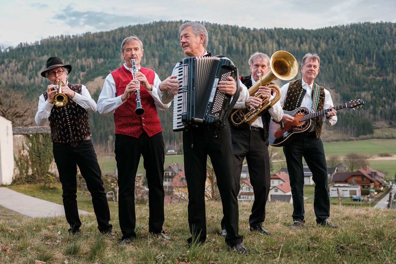 Fünf ältere Männer stehen in einem Grasfeld mit Bergen im Hintergrund und spielen jeweils ein anderes Instrument.