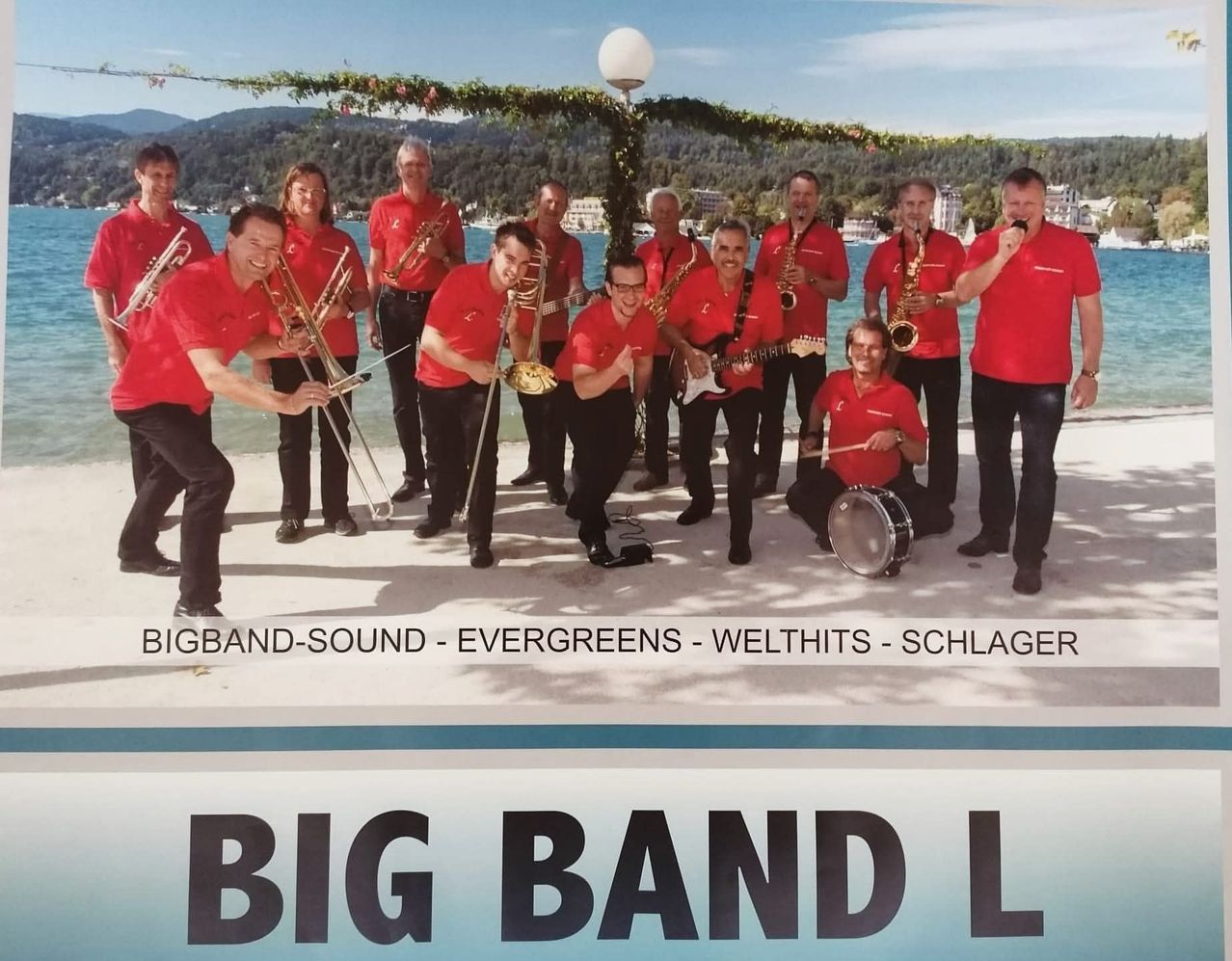 Eine Big Band von Musikern in roten Shirts und schwarzen Hosen, die verschiedene Instrumente halten, posiert für ein Foto am See. Der Text lautet 'BIGBAND-SOUND - EVERGREENS - WELTHITS - SCHLAGER'.