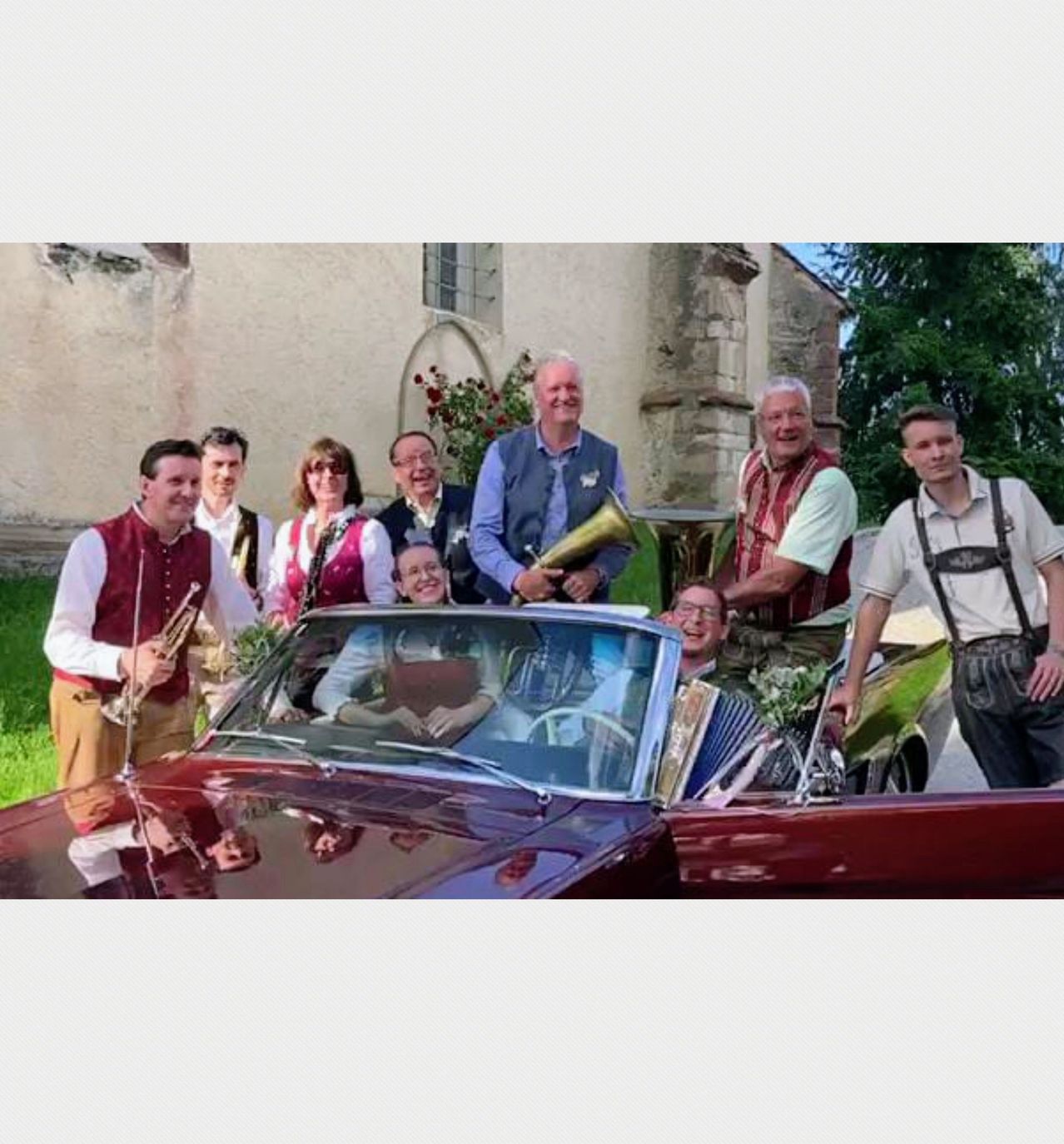 Eine Gruppe von Musikern steht neben einem roten Auto vor einem Gebäude mit alter Architektur. Sie halten Blechblasinstrumente.