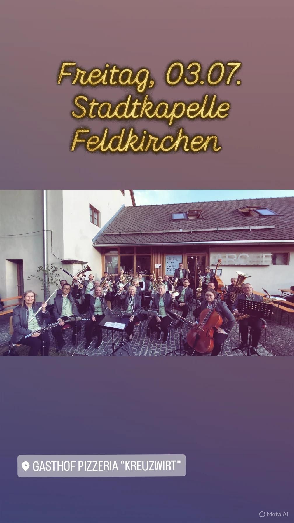 Eine Blechbläsergruppe steht vor einem Gebäude mit einem Schild, das Feldkirchen sagt. Die Musiker sitzen auf einem Kopfsteinpflaster.