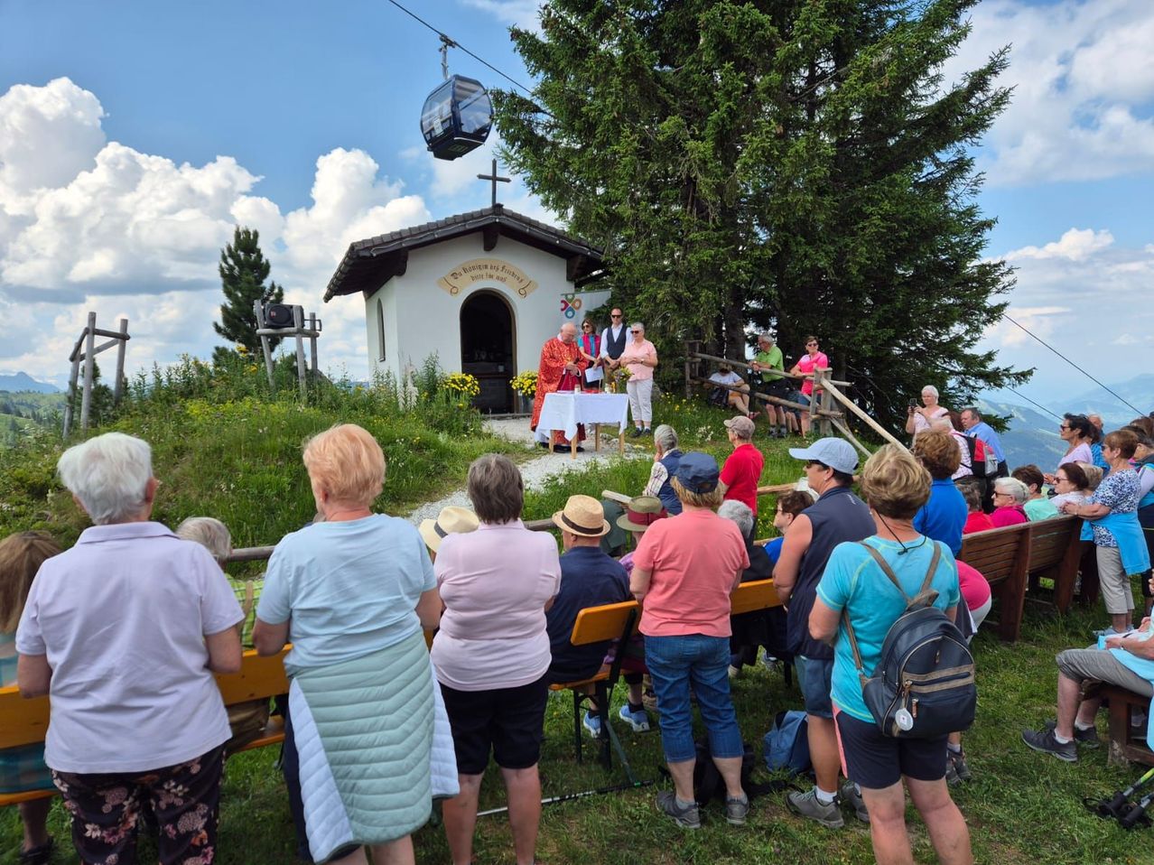 Eine Gruppe von Menschen versammelt sich vor einer kleinen Kapelle mit einem Kreuz auf dem Dach und beobachtet eine Zeremonie. Einige sitzen auf Bänken, andere stehen. Ein Tisch mit einer weißen Tischdecke ist aufgestellt.