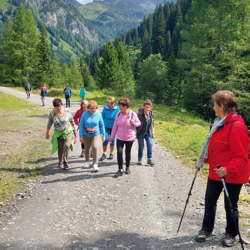 Eine Gruppe von Menschen, darunter Frauen, wandert auf einem Schotterweg in einem bergigen Gebiet. Sie tragen Turnschuhe und einige haben Rucksäcke.