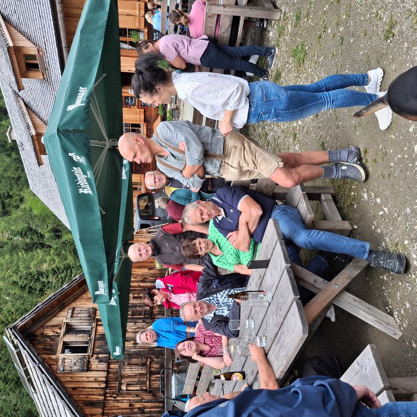 Eine Gruppe von Menschen sitzt und steht an Picknicktischen vor einem Holzgebäude mit einem grünen Schirm. Sie scheinen ihre Mahlzeit zu genießen.