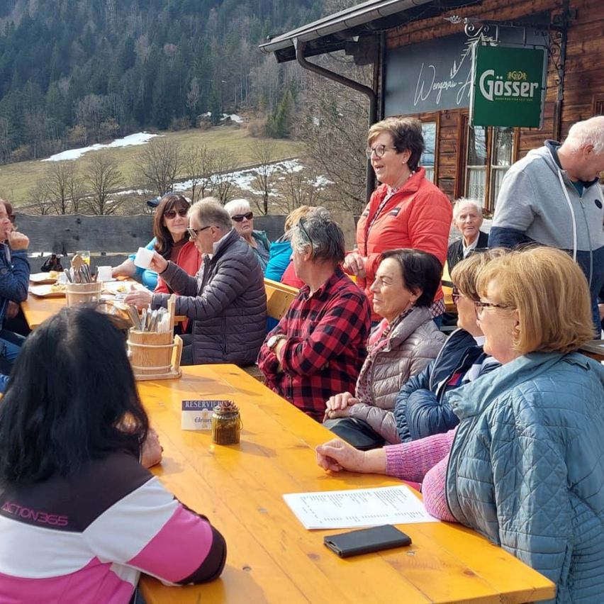 Mehrere Personen sitzen draußen an einem Holztisch und tragen Winterkleidung. Ein Schild für Gosser hängt am Gebäude. Bäume und Berge sind im Hintergrund.