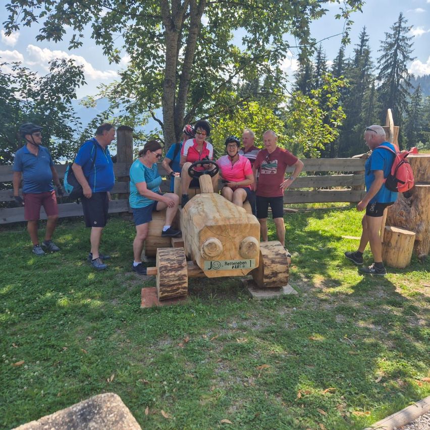 Eine Gruppe von Menschen steht um einen hölzernen Traktor auf einer Wiese mit einem Holzzaun und Bäumen im Hintergrund.