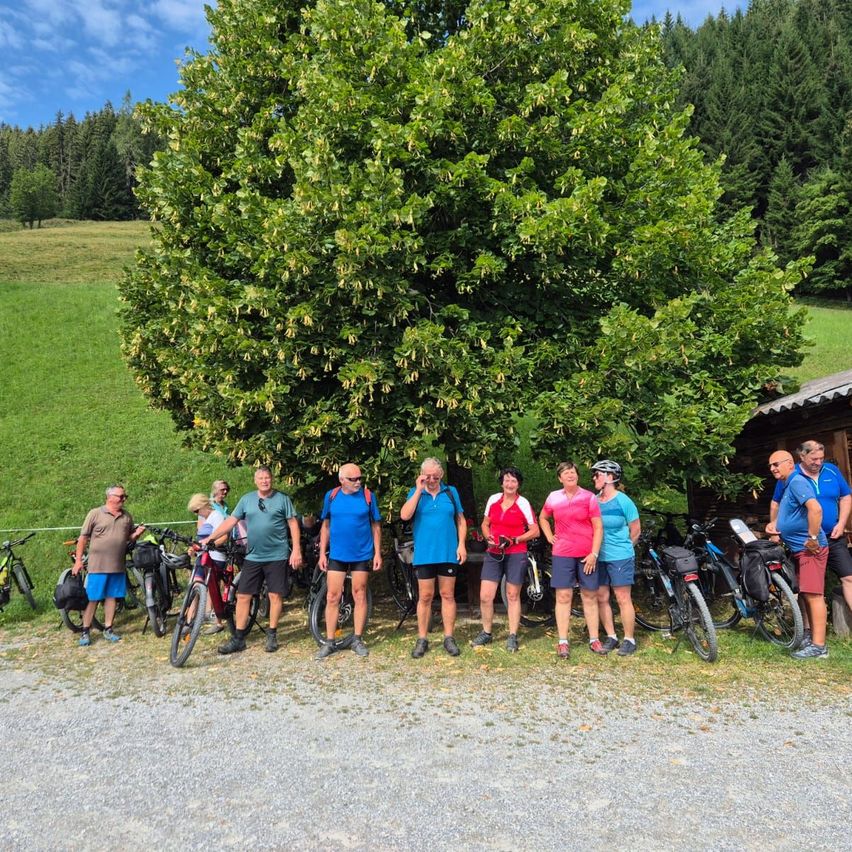 Eine Gruppe von Radfahrern, einige mit Helmen, stehen vor einem großen Baum in einem Grasfeld mit einem Holzgebäude im Hintergrund.