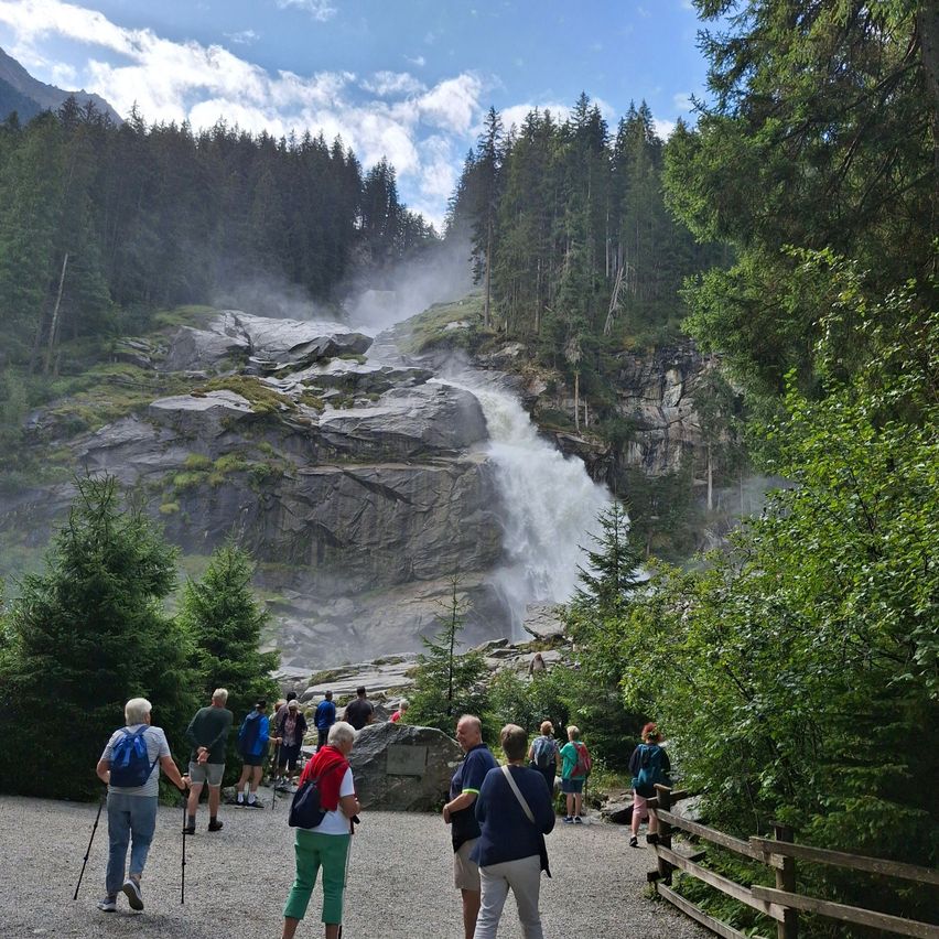 Eine Gruppe von Wanderern geht in der Nähe eines Wasserfalls auf einem Bergpfad. Der Wasserfall stürzt über felsige Klippen und ist von üppigem Grün umgeben.