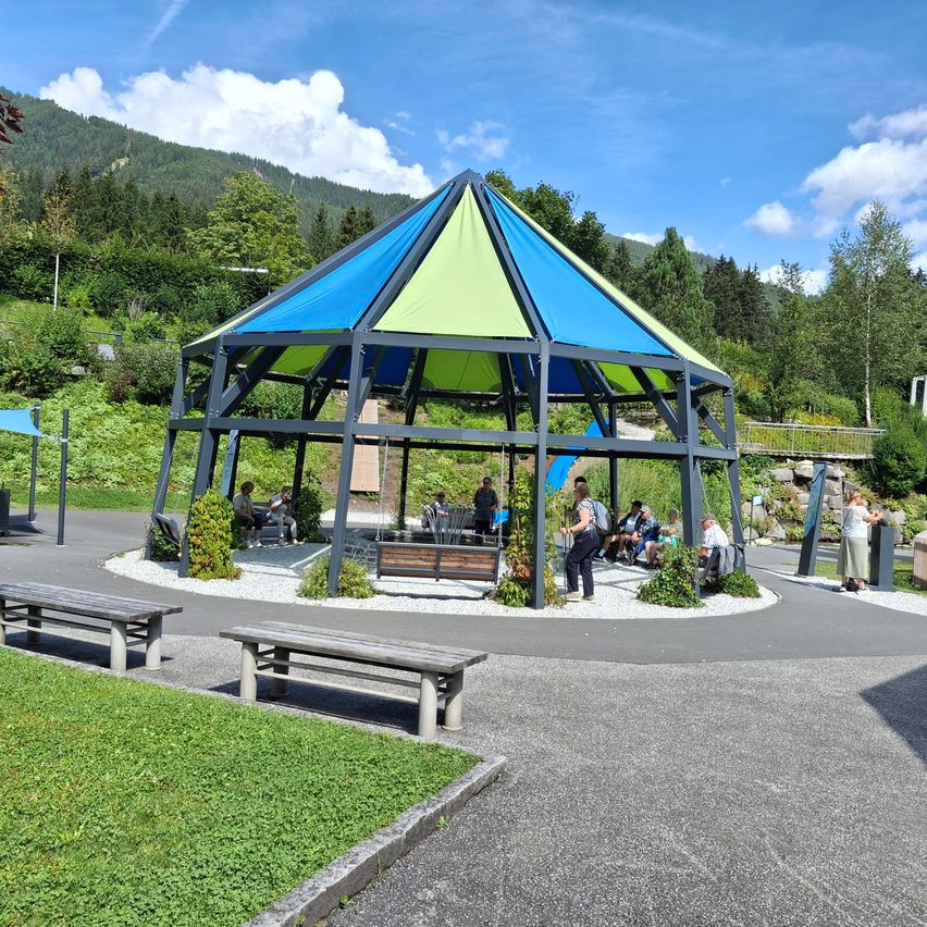Ein farbenfroher Pavillon mit blauem und grünem Dach ist von Bänken und Grün umgeben. Menschen sitzen und stehen darum herum und genießen die Aussicht auf die Berge.