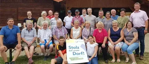 Eine Gruppe von Menschen posiert für ein Foto vor einem Holzhaus mit einem Schild Stolz auf unser Dorf! Sie sitzen auf Bänken und auf dem Gras.