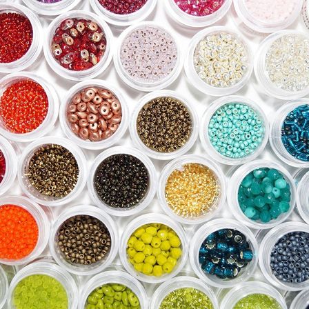 Eine Vielzahl von farbigen Perlen ist in durchsichtigen Plastikbehältern auf einer weißen Oberfläche angeordnet. Jeder Behälter enthält eine andere Farbe und Größe von Perlen, von rot bis grün und blau.