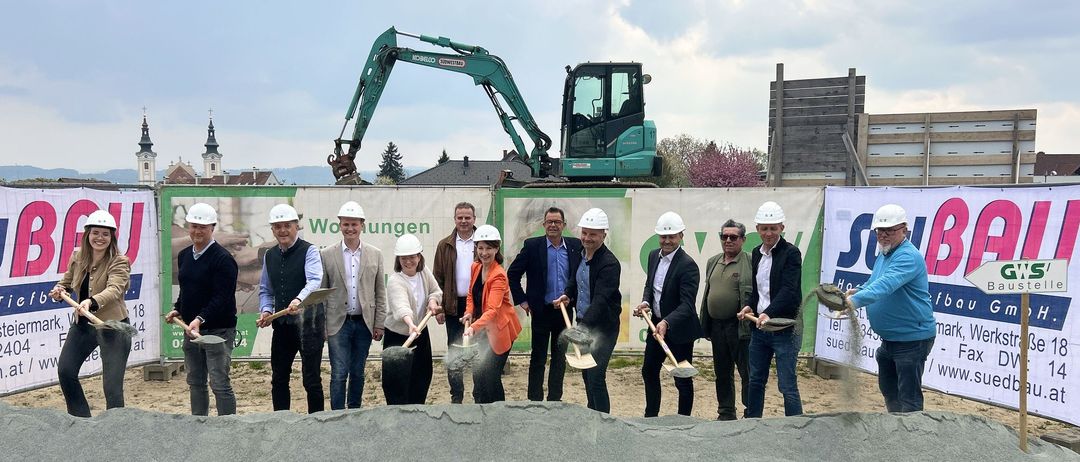 Eine Gruppe von Personen in Schutzhelmen hält Schaufeln über einer Sandaufschüttung, wahrscheinlich für eine Grundsteinlegungszeremonie. Hinter ihnen befindet sich ein Banner mit grünem Text, und ein grüner Bagger ist im Hintergrund zu sehen.