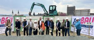 Eine Gruppe von Personen in Schutzhelmen hält Schaufeln über einer Sandaufschüttung, wahrscheinlich für eine Grundsteinlegungszeremonie. Hinter ihnen befindet sich ein Banner mit grünem Text, und ein grüner Bagger ist im Hintergrund zu sehen.