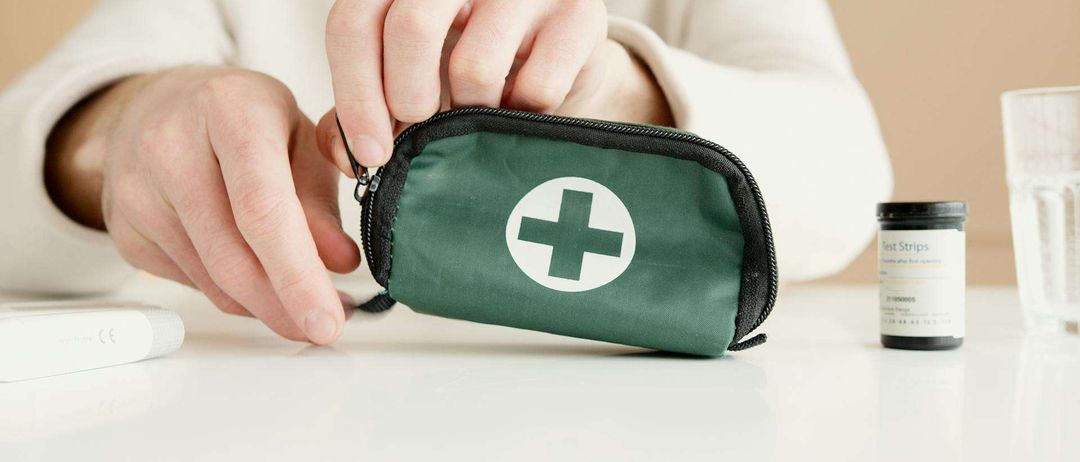 Eine Person hält eine grüne Erste-Hilfe-Tasche mit weißem Kreuzlogo auf einem Tisch.