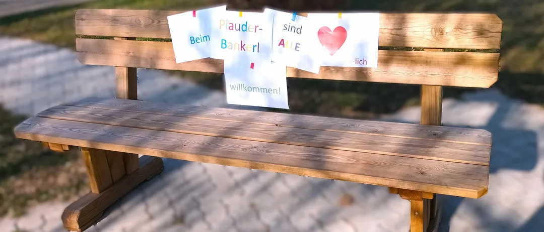 Eine Holzbank mit Schildern, die 'Plauder-Bankerl', 'Aile sind', '-lich' und 'willkommen!' besagen. Die Bank steht in einem Park. Unten befinden sich Logos von 'Gesundheit Osterreich GmbH' und 'Fonds Gesundes Osterreich'.
