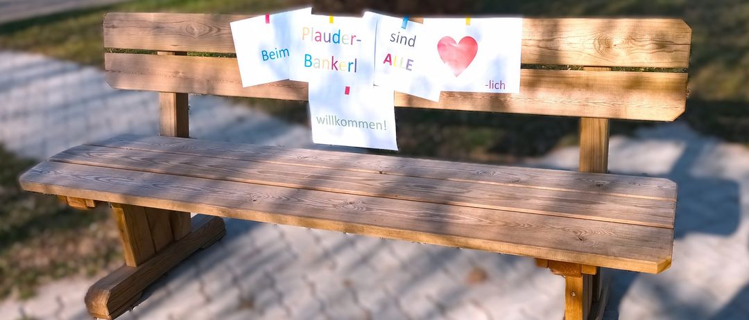 Eine Holzbank mit Schildern, die 'Plauder-Bankerl', 'Aile sind', '-lich' und 'willkommen!' besagen. Die Bank steht in einem Park. Unten befinden sich Logos von 'Gesundheit Osterreich GmbH' und 'Fonds Gesundes Osterreich'.