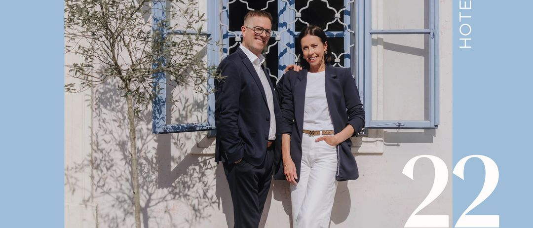 Zwei Menschen in formeller Kleidung stehen lächelnd vor einem Gebäude. Neben ihnen steht ein Baum, hinter ihnen befindet sich ein blaues Fenster und eine Lampe. Der Text lautet '22 Mai 26'.