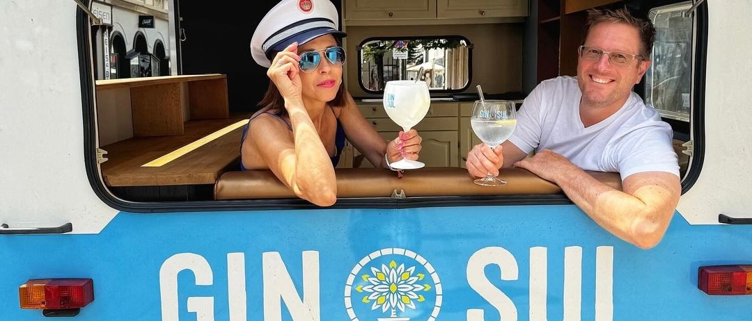 Ein Mann und eine Frau sitzen in einem Van mit offenen Fenstern, lächeln und halten Gläser mit Gin Tonic in der Hand. Der Van hat die Worte Gin Sul an seiner Seite.