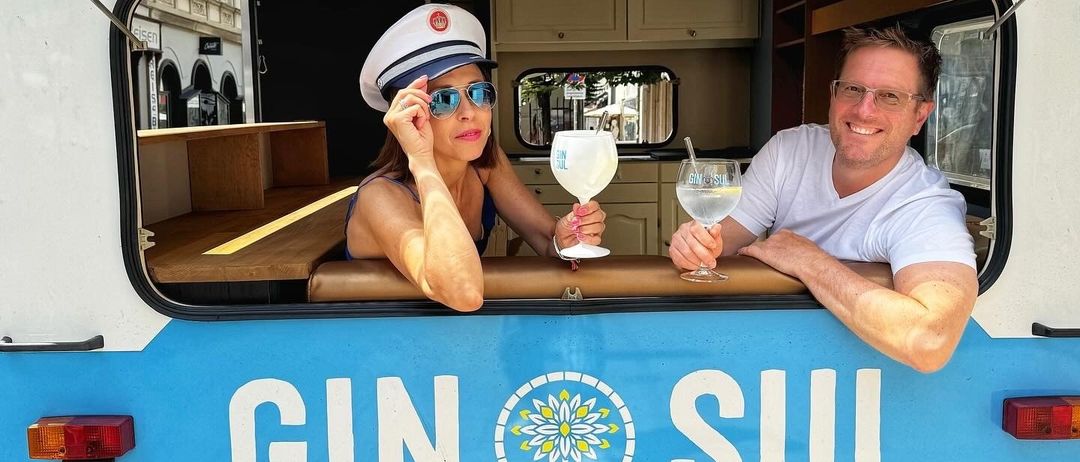 Ein Mann und eine Frau sitzen in einem blau-weißen Anhänger mit Gin Sul auf der Seite, halten Getränke in den Händen und lächeln.