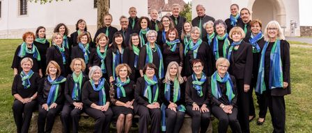 A choir group, dressed in black, poses for a photo in front of a building with a tree in the foreground.