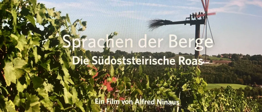 Eine Weinbergszene mit einem Windrad im Hintergrund, das einen klaren blauen Himmel und üppiges grünes Laub zeigt. Der Textüberlagerung lautet 'Sprachen der Berge' und nennt 'Ein Film von Alfred Ninaus.'