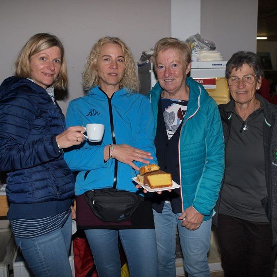 Vier Frauen stehen eng beieinander. Die Frau auf der linken Seite hält eine weiße Tasse. Die Frau in der Mitte hält einen Teller mit einem Stück Kuchen. Die Frau auf der rechten Seite trägt eine Brille.