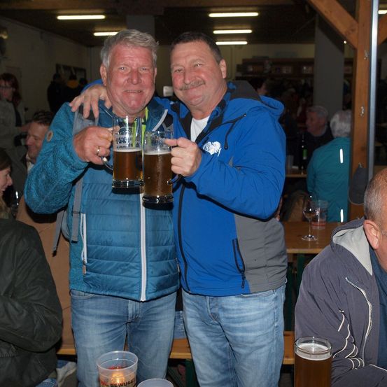 Zwei Männer in Jacken halten große Gläser Bier, lächeln und posieren für ein Foto in einem überfüllten Raum. Hinter ihnen sitzen und stehen Leute, einige halten Getränke.