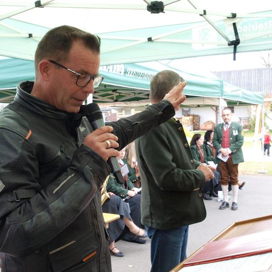 Ein Mann spricht in ein Mikrofon bei einer Veranstaltung im Freien, unter einem Zelt, mit anderen, die in traditioneller deutscher Kleidung im Hintergrund stehen.