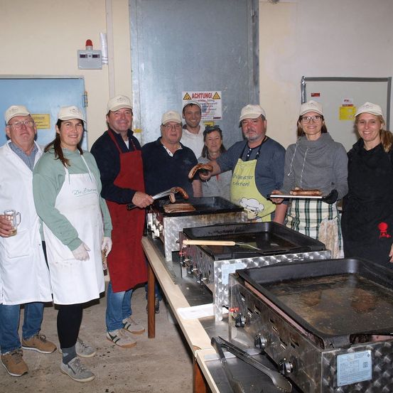 Eine Gruppe von Menschen in Schürzen und Mützen kocht Würste auf Grills in einem Raum. Eine Person hält eine Schale mit Würsten. Ein Warnschild ist an der Wand angebracht.