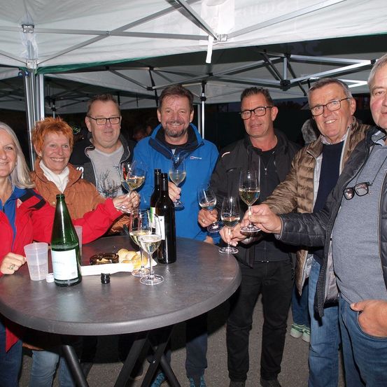 Eine Gruppe von Menschen, sowohl Männer als auch Frauen, versammelt sich um einen Tisch unter einem Zelt und hält Weingläser. Der Tisch hat Weinflaschen, Gläser und etwas Essen. Sie scheinen ein Ereignis zu feiern.