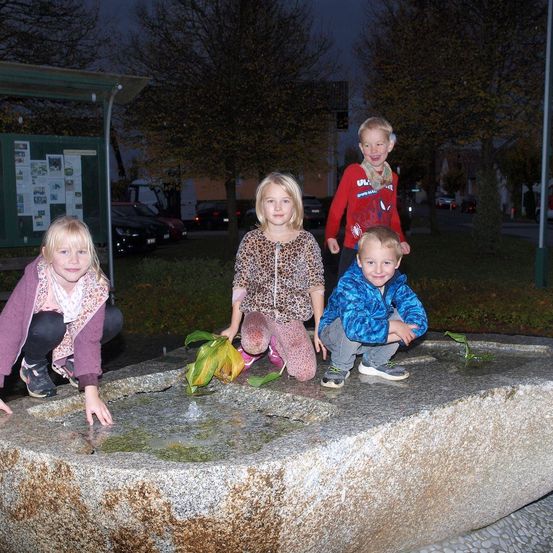 Vier Kinder spielen nachts mit Wasser in einem Brunnen. Sie sind in Winterkleidung gekleidet. Dahinter befinden sich Bäume, eine Bushaltestelle und geparkte Autos.