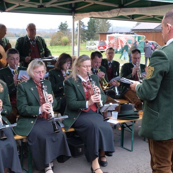 Eine Freiluftveranstaltung mit einer Blaskapelle, die Instrumente spielt. Einige Bandmitglieder sitzen auf einer Bank, während andere stehen. Das Publikum sitzt unter einem Zelt.