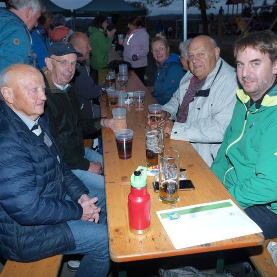 Vier Männer sitzen um einen Tisch herum, lächeln und haben Getränke vor sich. Einer hält ein Glas Bier. Ein weiterer Mann steht hinter ihnen.