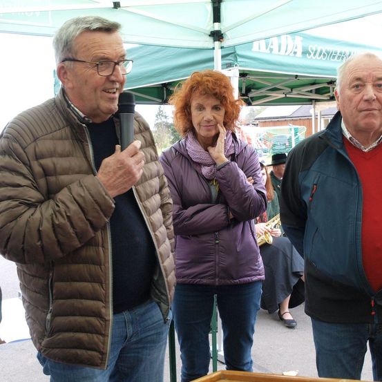 Drei Erwachsene, zwei Männer und eine Frau, stehen vor einem Zelt. Der Mann auf der linken Seite hält ein Mikrofon. Die Frau in der Mitte hat die Arme verschränkt. Dahinter steht ein Zelt mit der Aufschrift 'ADA'.