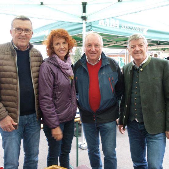Vier Personen stehen unter einem Zelt mit der Aufschrift KADA SUSSTEWARK. Ein Mann in einer braunen Jacke, eine Frau in einer lila Jacke, ein Mann in einem roten Hemd und ein Mann in einer grünen Jacke lächeln alle.
