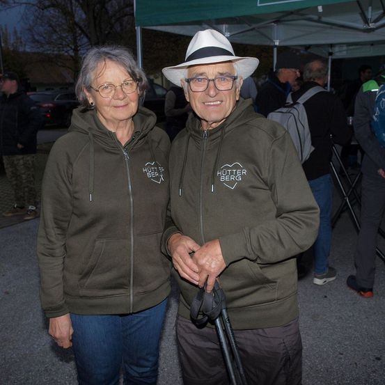 Zwei Menschen, ein älterer Mann und eine ältere Frau, stehen nebeneinander in grünen Hoodies mit weißen Logos. Sie lächeln und halten Stöcke. Sie stehen unter einem grünen Zelt.