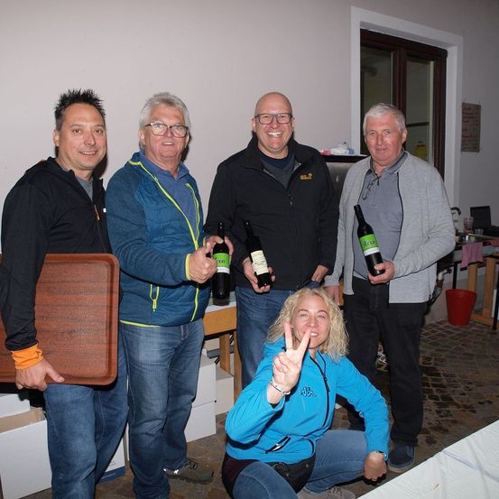 Fünf Erwachsene, vier Männer und eine Frau, posieren für ein Foto. Sie alle lächeln und halten Weinflaschen. Der Mann in der Mitte hält zwei Flaschen. Die Frau kniet und macht ein Friedenszeichen. Hinter ihnen steht ein Holztisch mit einigen Gegenständen darauf.