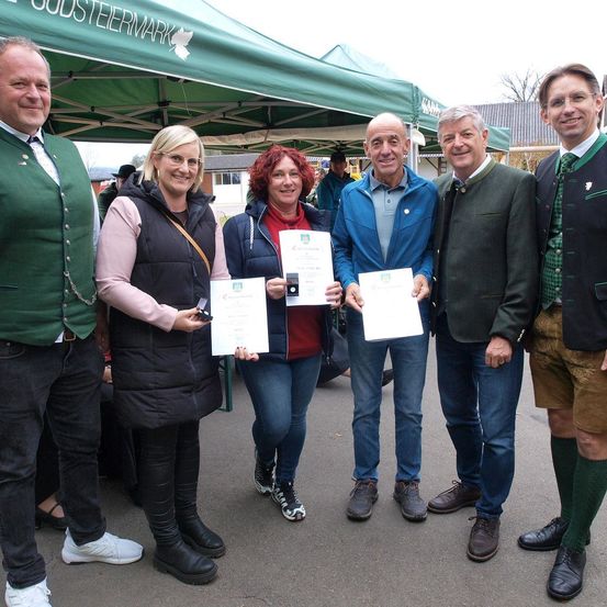 Fünf Personen stehen unter einem grünen Zelt mit einem Logo und halten Urkunden in den Händen. Sie scheinen sich bei einer Veranstaltung zu befinden.