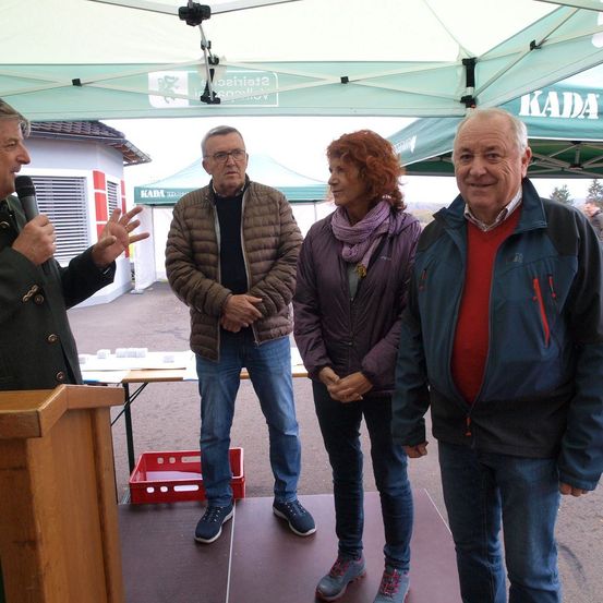 Vier Menschen stehen unter einem Zelt, zwei Männer und zwei Frauen. Ein Mann spricht in ein Mikrofon, während die anderen drei zuhören. Hinter ihnen steht ein Tisch mit Papieren und einem roten Korb.