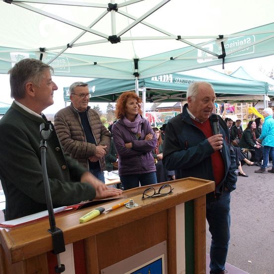Vier Menschen stehen in der Nähe eines Podiums im Freien. Ein Mann spricht in ein Mikrofon, während ein anderer einen Marker hält. Dahinter stehen zwei Frauen mit verschränkten Armen. Mehrere Menschen sitzen auf der Wiese, und ein Zelt ist in der Nähe.