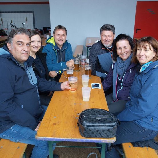 Eine Gruppe von Menschen sitzt an einem Tisch mit Biergläsern und lächelt für ein Foto. Sie tragen Jacken und einige Schals. Eine Handtasche liegt auf dem Tisch, und der Raum hat Holzbänke und eine rote Tür.